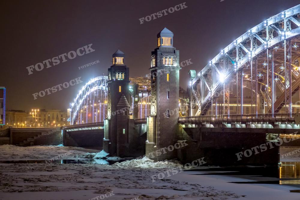 На фотографии ночной вид на Большеохтинский мост в Санкт-Петербурге, освещённый яркими огнями. Мост и его отражение в реке, частично покрытой льдом, создают живописную картину. Изображение подходит для иллюстрирования городской ночной жизни, статей о зимнем периоде в городе и может быть использовано в рекламных целях для привлечения туристов.